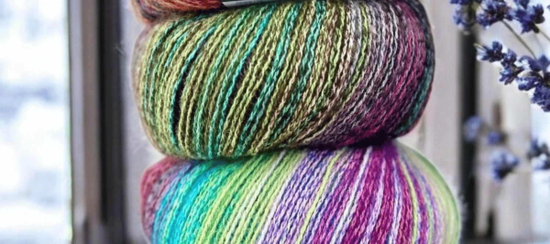 Two colorful balls of variegated yarn stacked on top of each other near a window, with purple flowers visible on the right side of the image.