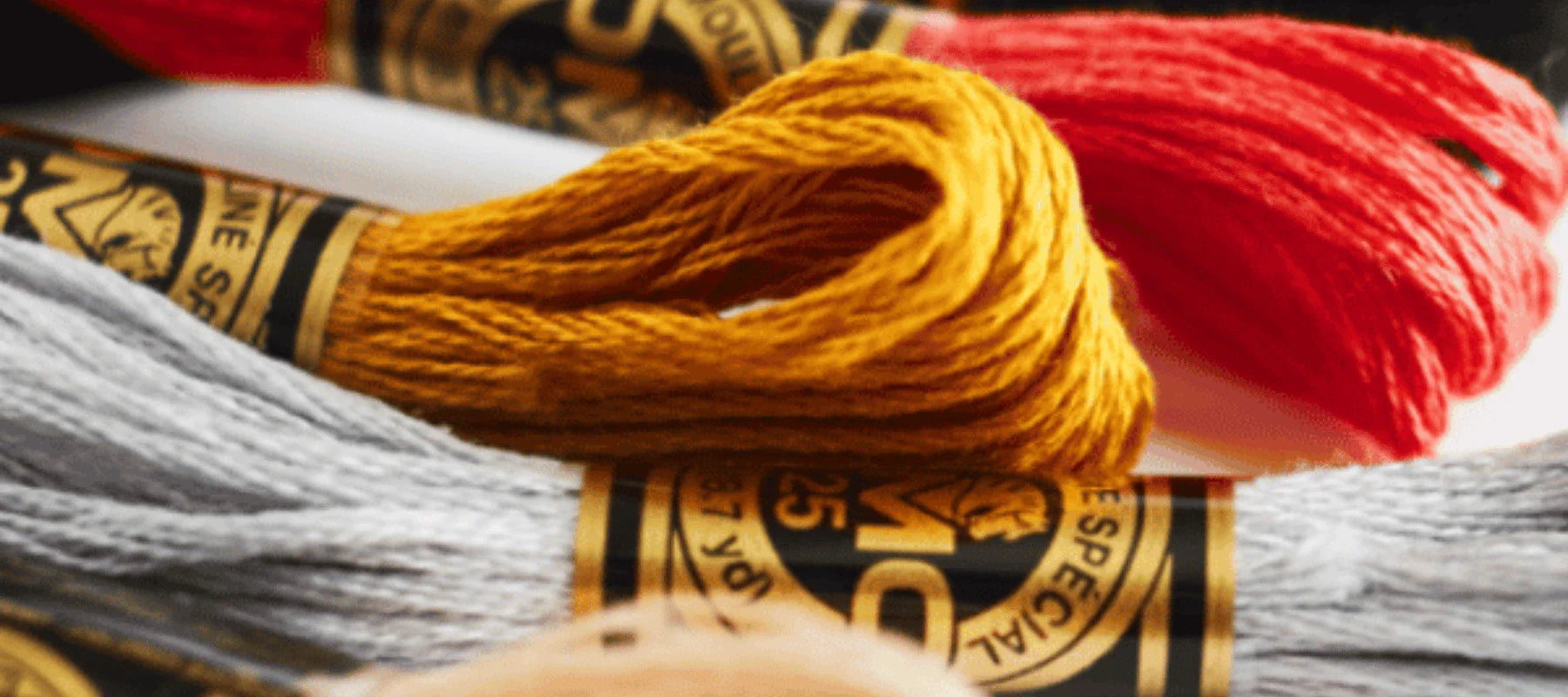 Close-up of embroidery floss skeins in gray, yellow, and red, each wrapped with a black and gold label showing the number 25.