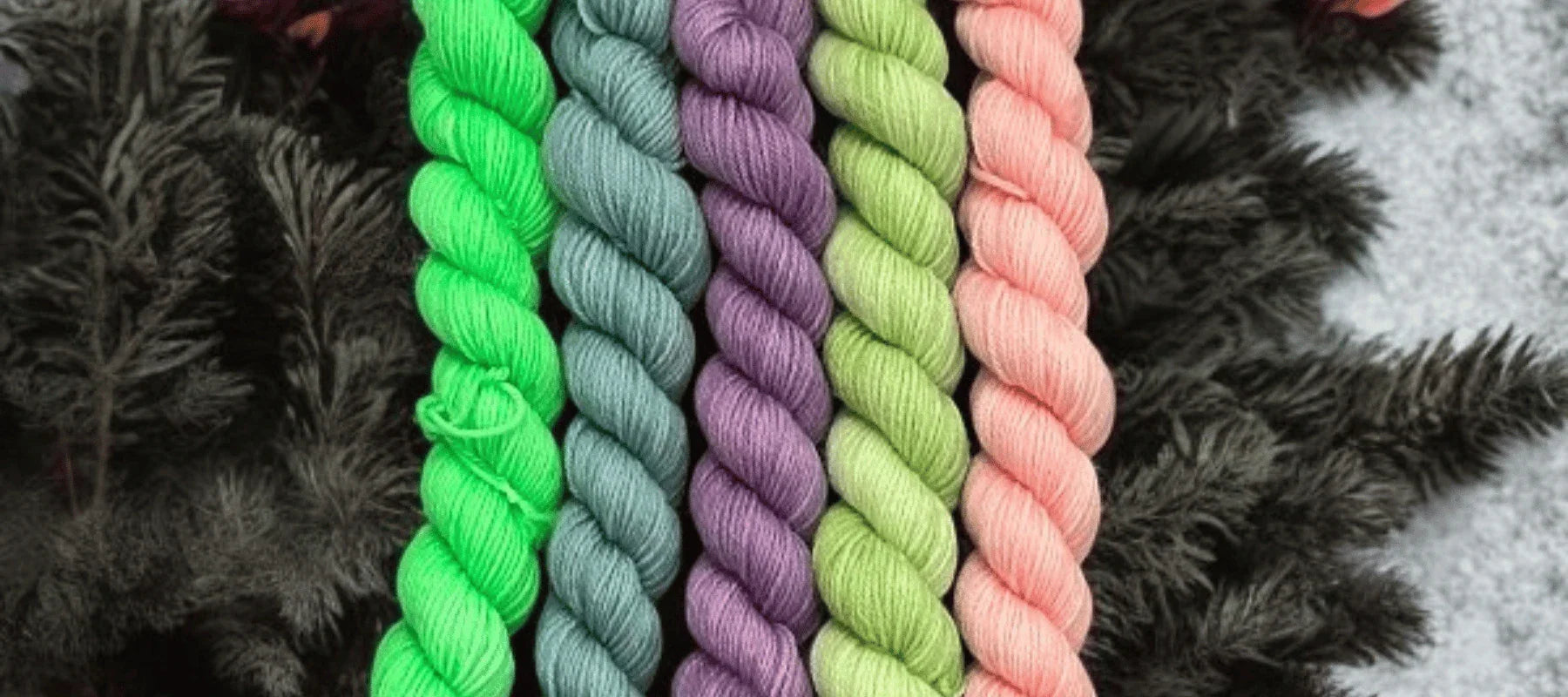Six skeins of yarn in bright green, gray, purple, light green, peach, and pink shades are neatly arranged in a row on a dark, textured background.