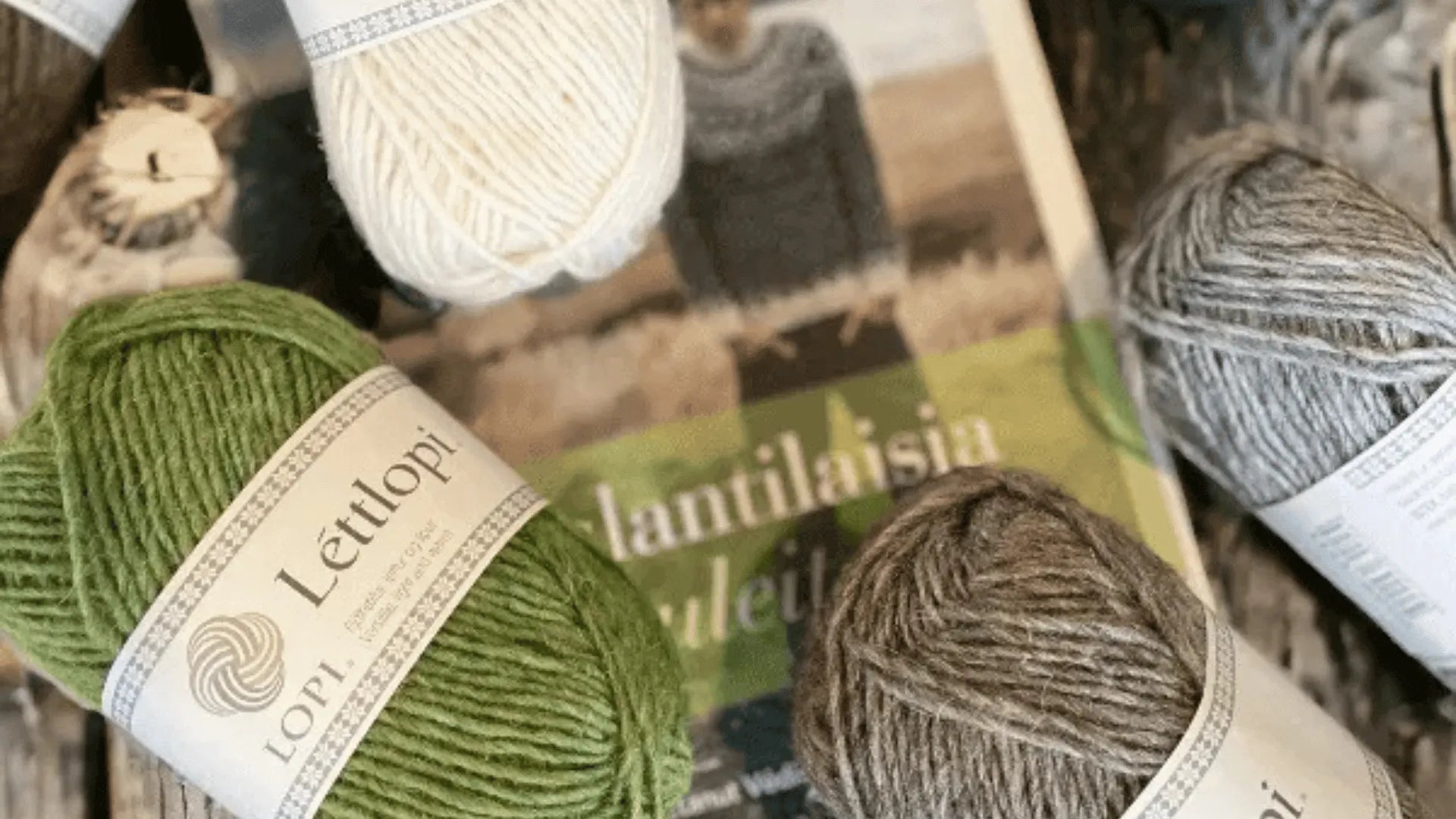 Four skeins of Lopi yarn in green, white, brown, and gray are arranged around a knitting book with a blurred cover featuring a person wearing a sweater.