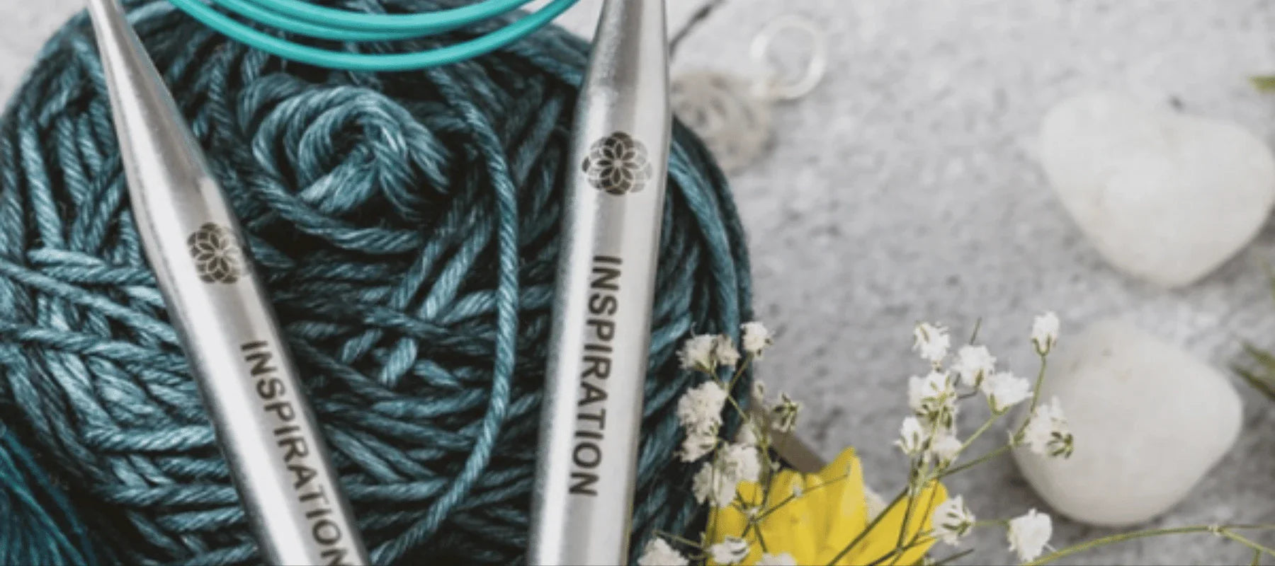 Two metal knitting needles labeled “INSPIRATION” rest on a ball of teal yarn, surrounded by small white flowers, yellow leaves, and white stones on a gray surface.