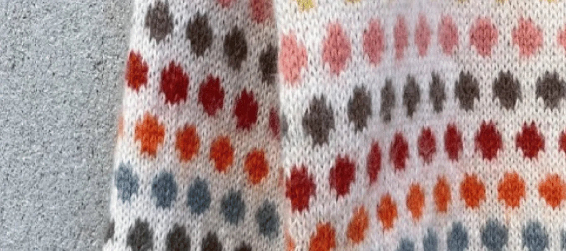 A close-up of knitted fabric with rows of colorful dots in blue, orange, red, and brown on a white background, next to a textured gray surface.