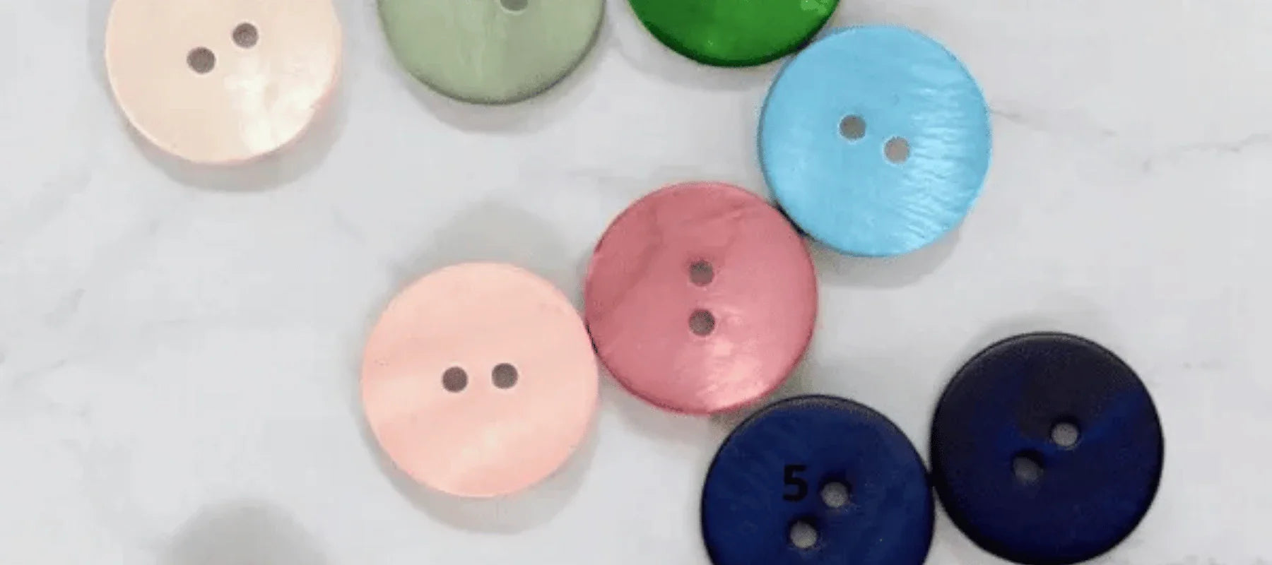 Seven colorful, round buttons with two holes each are scattered on a white surface. Button colors include pink, green, blue, navy, and light gray. One navy button is labeled with the number 5.