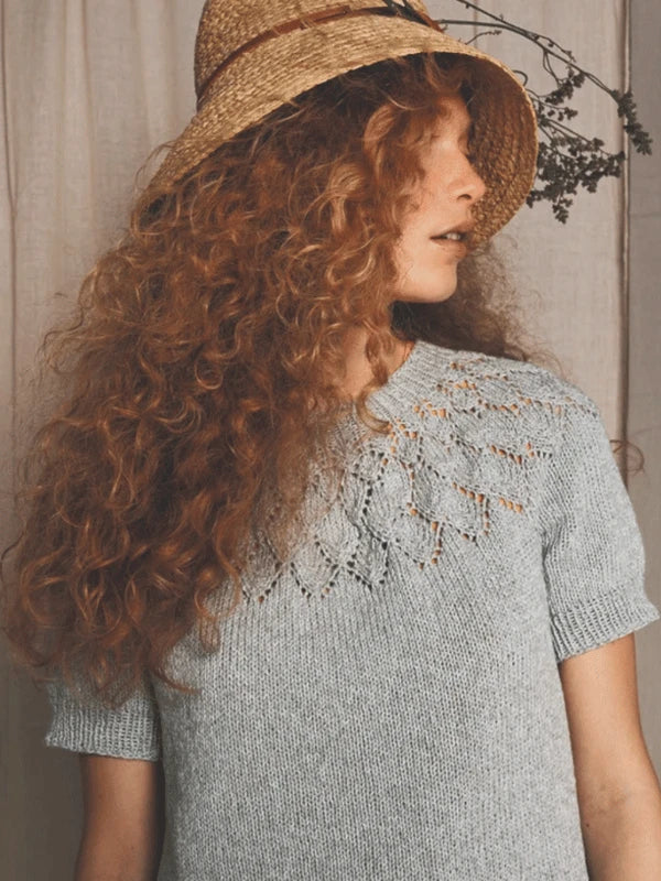 Woman with long curly red hair wearing a straw hat and a light grey knitted top with a lacy pattern at the neckline, looking to the side against a neutral background.
