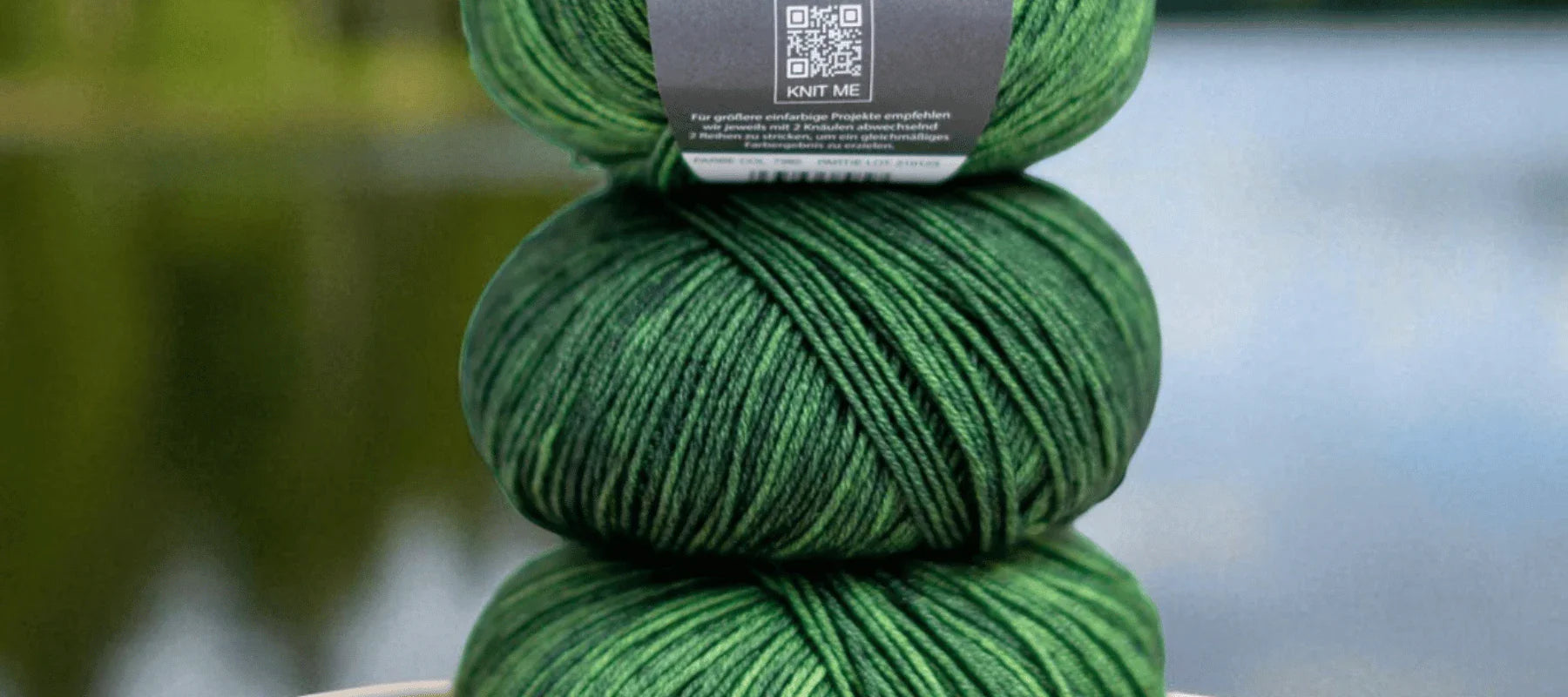 Three skeins of green yarn, stacked vertically with a label featuring a QR code on the top skein, are set against a blurred natural background.