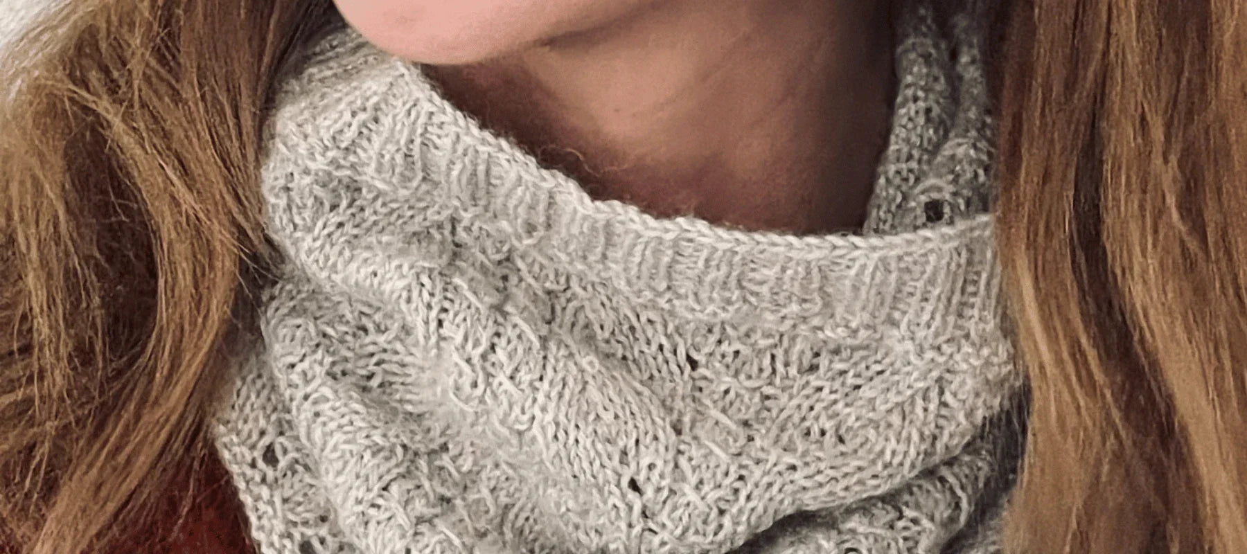 Close-up of a person wearing a light gray, textured knit scarf around their neck. The person has long, light brown hair and is dressed in a reddish top.