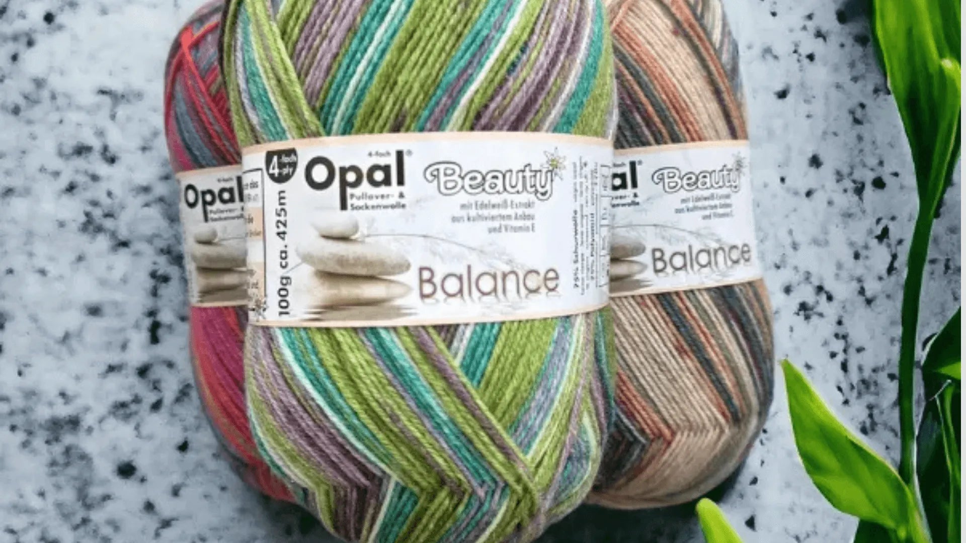 Three skeins of Opal Beauty Balance yarn in multicolored stripes are stacked together against a speckled gray background, with green leaves visible on the right side.