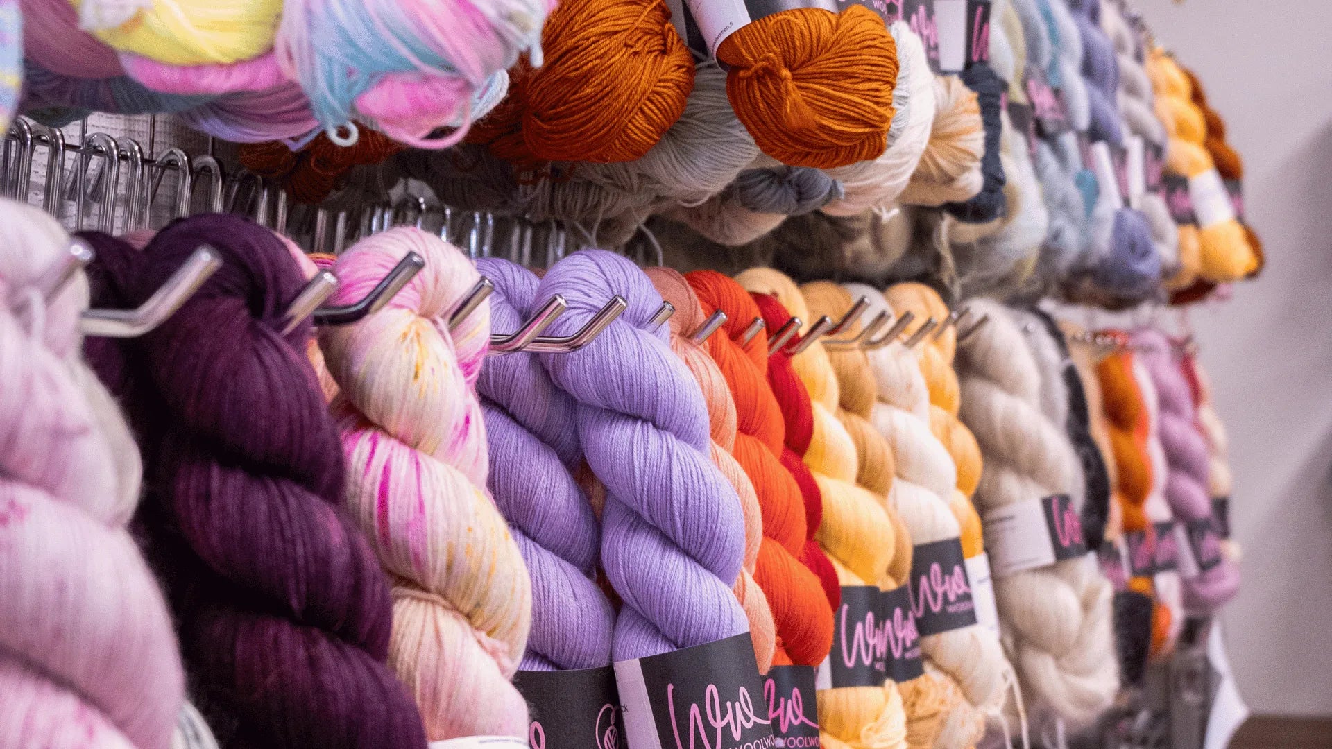 Colorful skeins of yarn in various shades, including purple, yellow, orange, cream, and pastel hues, neatly displayed on metal racks in a yarn or craft store.