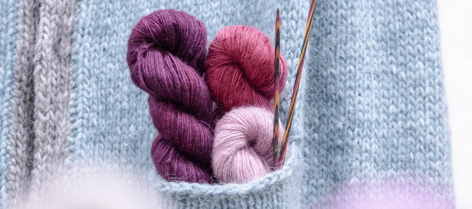 Three skeins of purple, red, and light pink yarn and several knitting needles are tucked into the pocket of a light blue knitted garment.