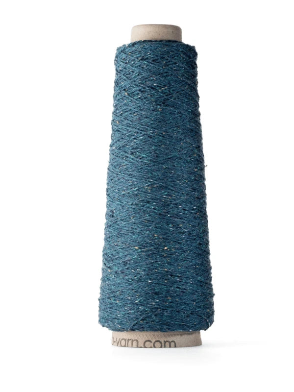A tall spool of textured teal-blue Ito Kinu silk yarn with subtle lighter flecks, wound on a cardboard cone and standing upright on a white background—perfect for kesäneuleisiin projects.