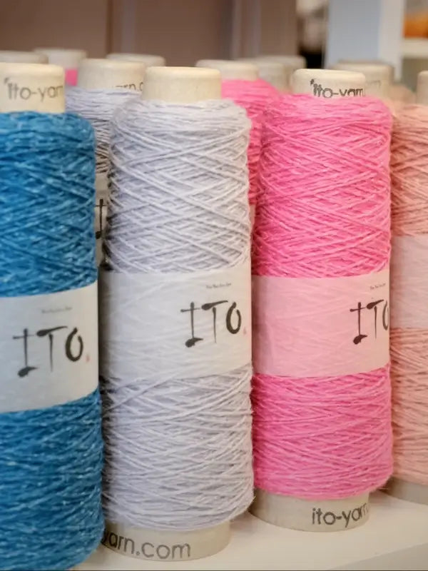 Several large spools of yarn in blue, gray, and pink are standing upright in a row. Each spool features a white label with “ITO” and “ito-yarn.com” at the bottom edge. Among them is the luxurious Ito Yarn Shimo, crafted from kierrätetty silkki (recycled silk) and merinovillalanka (merino wool yarn), combining sustainability and softness for your next project.
