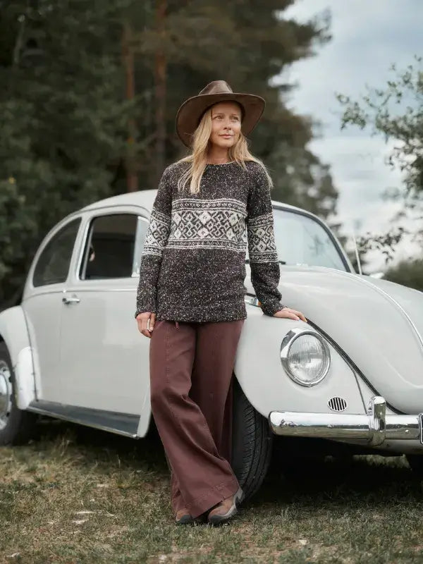 A woman wearing a Jenna Kostet - Neulottu Kalevala III, Ennakkotilaus knit sweater with a dark kirjoneule pattern and a wide-brimmed hat leans against a light-colored vintage car.