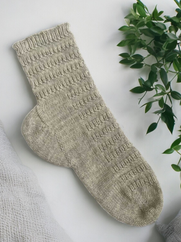 A pair of light gray socks made with the Juhani sukkaohje PDF and Lang Super Soxx 6ply yarn are laid flat on a white surface beside a green leafy plant and the corner of gray knitted fabric.