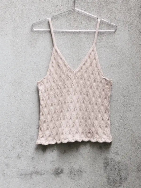 The Knitting For Olive Olive Top Neuleohje + Pure Silk is a light beige, sleeveless knitted top with a scalloped hem and diamond pattern, shown on a clear hanger against a textured gray wall.