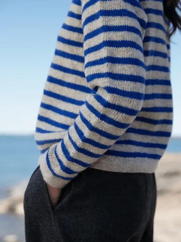 A person in a blue and gray striped sweater and dark pants stands by the water under a clear sky, reflecting the pohjoismainen estetiikka showcased in Laine Juhlanumero - ENNAKKO.