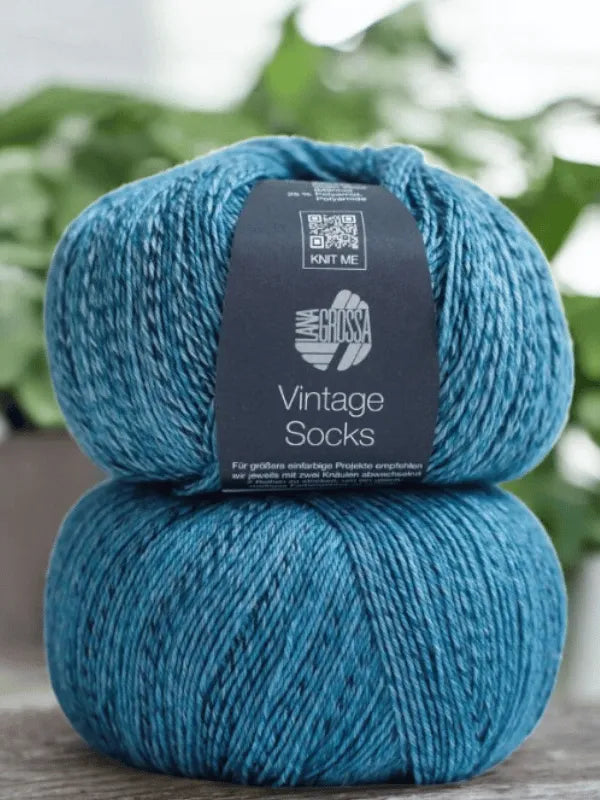 Two skeins of blue marled yarn are stacked, the top labeled "Lauri Fingerin sukkaohje + Lana Grossa Vintage socks"—ideal for knitting villasukat. Blurred green plants can be seen in the background.