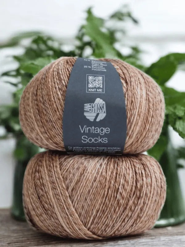 Two balls of Lana Grossa Vintage socks yarn are stacked on wood with a blurred green background, ideal for knitting villasukat or using the Lauri Fingerin sukkaohje +Lana Grossa Vintage socks pattern.