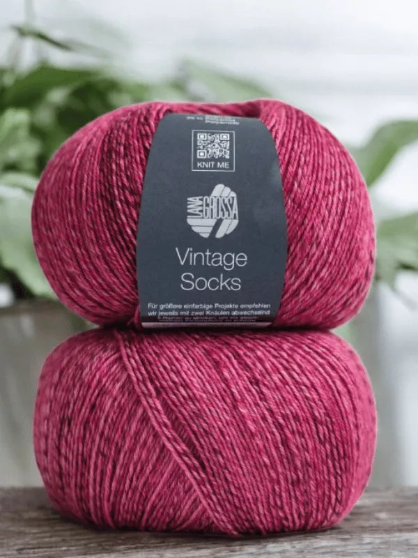 Two skeins of Lana Grossa Vintage Socks red-pink yarn are stacked, labeled “Vintage Socks,” with soft green leaves in the background. Great for villasukat or pairing with your Lauri Fingerin sukkaohje +Lana Grossa Vintage socks pattern.