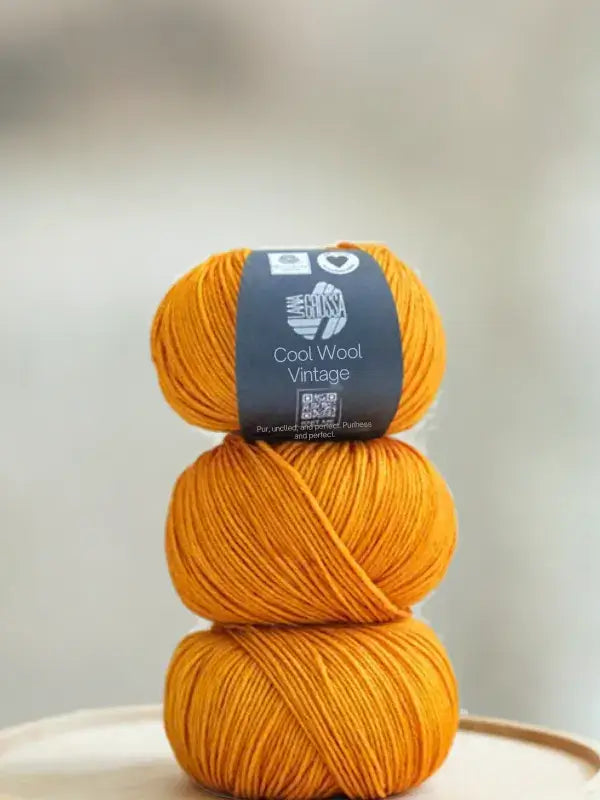 A vertical stack of three skeins of vibrant orange Lana Grossa Cool wool vintage yarn, with a label around the top skein.