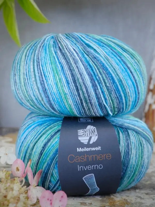 Two skeins of Lana Grossa Meilenweit 100 Cashmere Inverno yarn in variegated shades of blue, green, and white are stacked together, their label clearly visible. This luxurious sukkalanka features soft cashmere fibers for added comfort. Nearby, flowers and a green leaf accentuate the display.