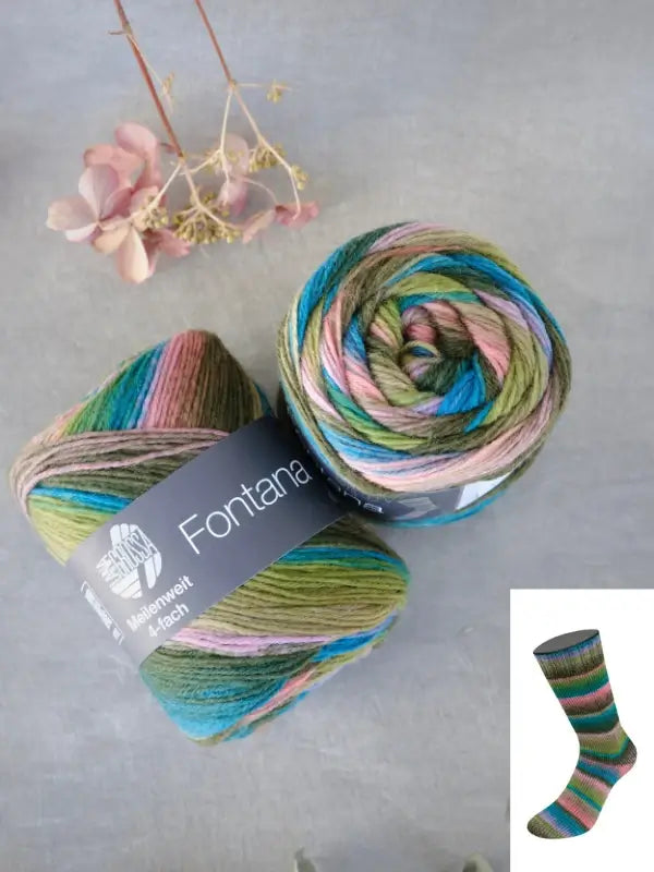 Two skeins of Lana Grossa Meilenweit 100 Fontana yarn in multicolored shades of green, blue, pink, and brown rest on a gray surface with dried flowers nearby. An inset displays a knitted sock crafted from the same Lana Grossa Meilenweit 100 Fontana yarn.