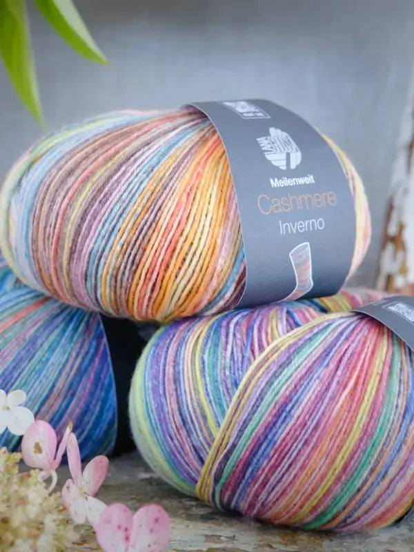 Three skeins of Lana Grossa Meilenweit 100 Cashmere Inverno multicolored yarn are stacked together, displaying soft pastel hues and pink flowers in the foreground—ideal for crafting delicate sukkalanka projects with a luxurious touch of cashmere softness.