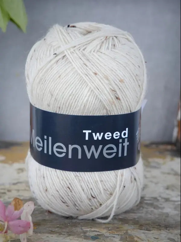 A ball of off-white Lana Grossa Meilenweit 100 Tweed yarn with small brown flecks, labeled Meilenweit Tweed, rests on a rustic wooden surface with a pink flower in the corner.
