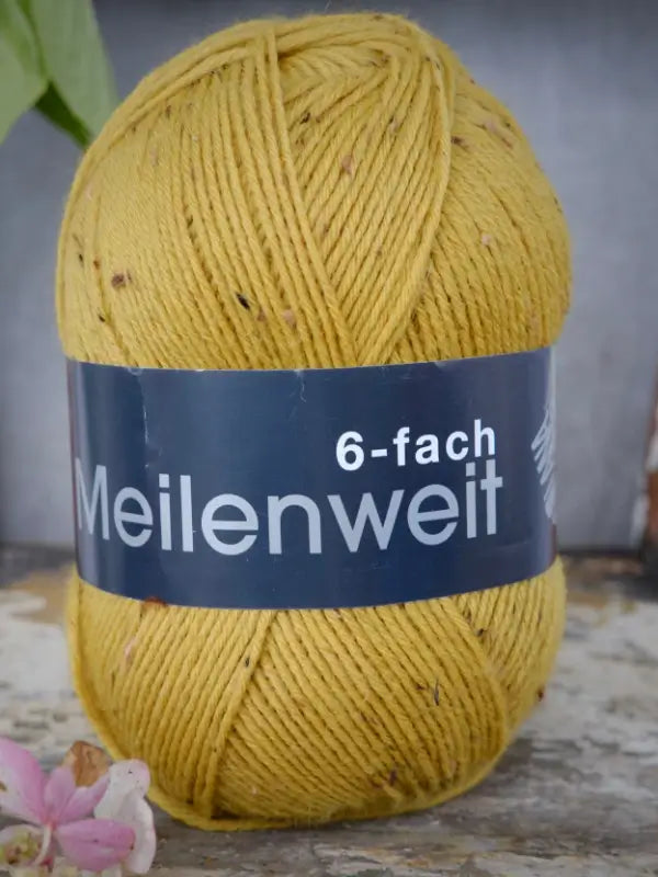 A skein of Lana Grossa Meilenweit 6 Tweed, a yellow speckled yarn with its recognizable label, rests on a rustic wooden surface beside a delicate pink flower.