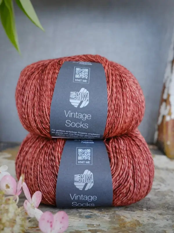 Two skeins of reddish-brown Lana Grossa Vintage Socks yarn are stacked on a rustic surface, accented by a few pink flowers and green leaves nearby. This soft merinosukkalanka adds a touch of timeless elegance to any knitting project.