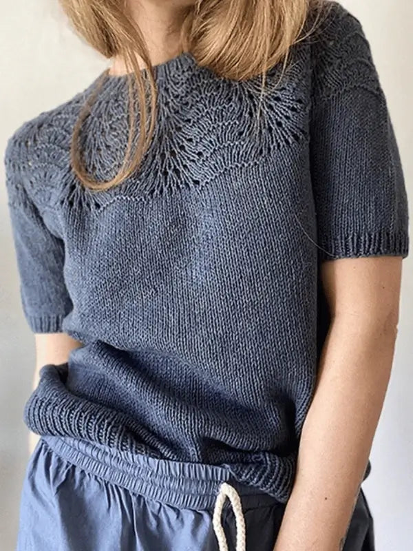 A person wears the Le Knit Peacock tee neuleohje, ENG—a short-sleeved, knitted blue top with a lace yoke pattern—paired with blue drawstring pants. Their hand is in their pocket and long hair partly covers their face.