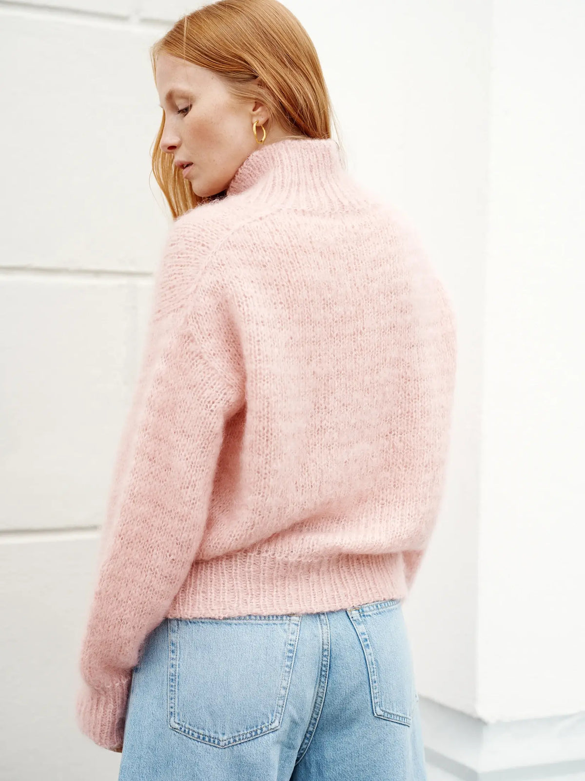 A person with long red hair embodies true Nordic Chic style, standing sideways and looking down against a white background while wearing light blue jeans and a pink fuzzy turtleneck sweater—perfectly capturing the timeless aesthetic of Leeni Hoimela’s “Skandityyli, Ajattomia ohjeita nykyneulojalle.”.
