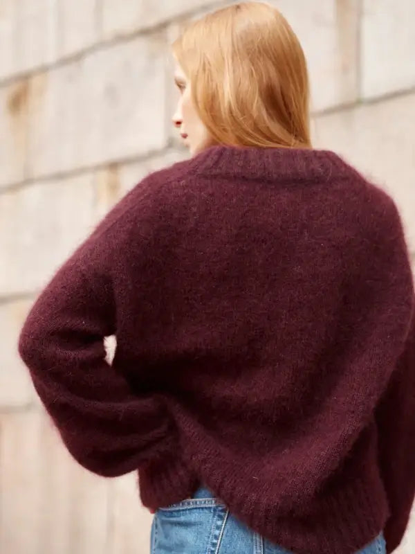 A person with long, light hair is seen from the back, dressed in a fuzzy burgundy sweater and blue jeans, standing outdoors near a stone wall—perfectly embodying the Nordic chic style featured in Leeni Hoimela’s *Skandityyli, Ajattomia ohjeita nykyneulojalle.*.