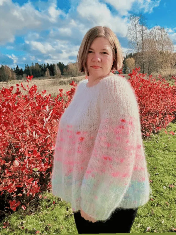 A woman with short brown hair stands outside among red bushes and green grass, wearing the Lempi pusero neuleohje—an oversized pastel sweater with pink and blue accents. Trees and a partly cloudy sky complete the background.