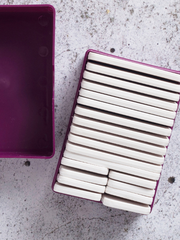 A purple rectangular container, partially open, holds neatly stacked white chewing gum pieces on a light speckled surface, arranged with precision reminiscent of KnitPron pingotuskammat.