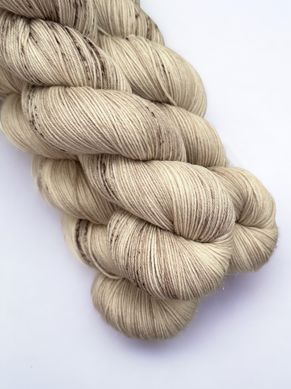 Two skeins of WoolWomen Merino Sock in light beige, hand-dyed with subtle brown speckles, are neatly twisted and placed on a plain white background.