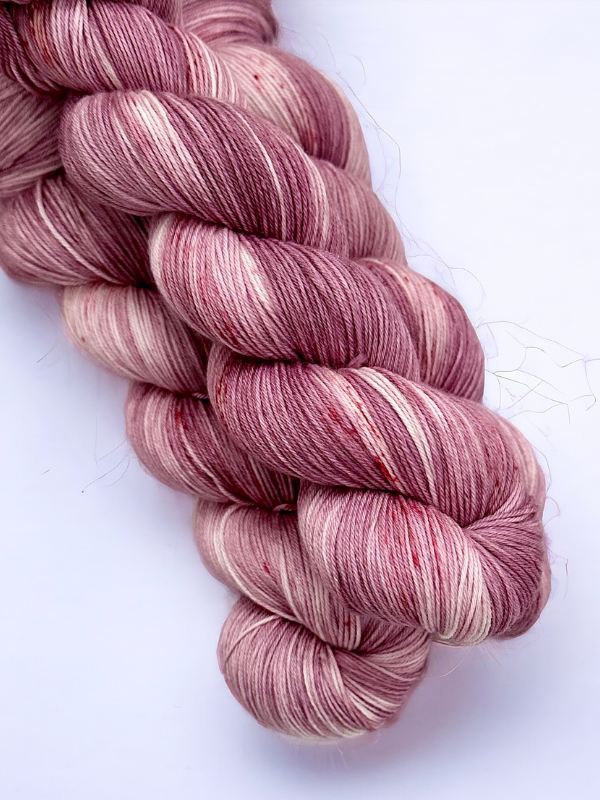 A close-up of a twisted skein of WoolWomen Merino Sock yarn in soft mauve, pink, and cream tones on a white background. This superwash merinovilla yarn is smooth with subtle color variations and a few visible specks.