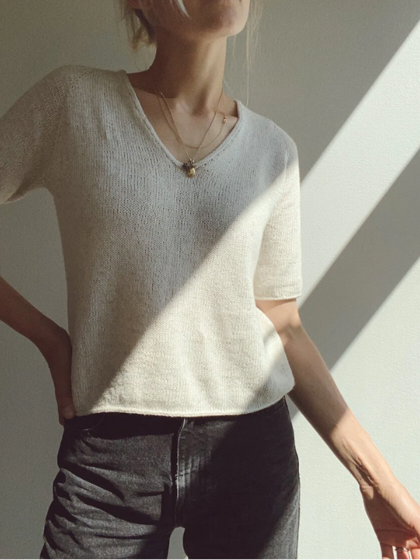 A person stands in sunlight wearing the PetiteKnit Cumulus Tee neuleohje, paired with dark pants and layered necklaces. Their face is mostly out of frame, hand on hip, as angular sunlight and shadows play across the wall.