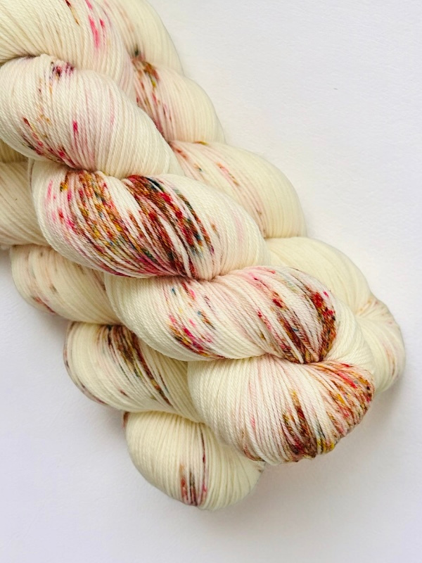 Three skeins of WoolWomen Merino Sock yarn in cream, speckled with brown, pink, and yellow, are neatly stacked against a white background. This superwash merino wool is perfect for your next project.