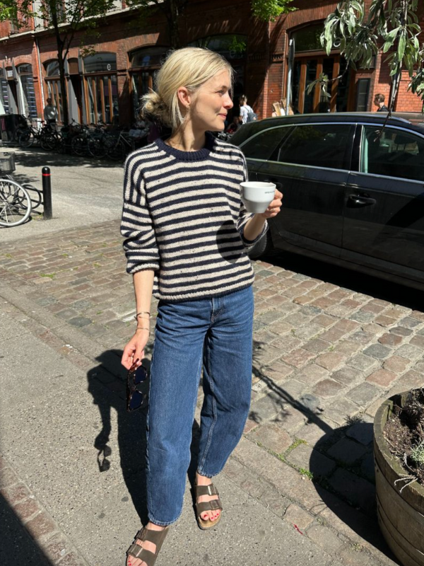 A blonde woman in a PetiteKnit Olga sweater neuleohje—black and white striped—blue jeans, and sandals stands on a sunny city sidewalk holding a white cup, with parked cars and shops in the background.