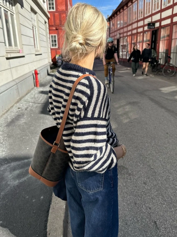 A blonde-haired person in a low bun wears blue jeans and a PetiteKnit Olga sweater neuleohje, standing on a street with a black shoulder bag as cyclists and pedestrians pass red and grey buildings.