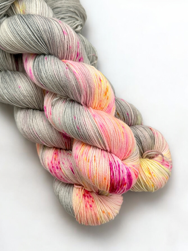 Two skeins of WoolWomen Merino Sock are intertwined on a white background. The superwash merino yarn is mostly gray with soft pink, yellow, and specks of bright pink and orange for a vibrant gradient effect.