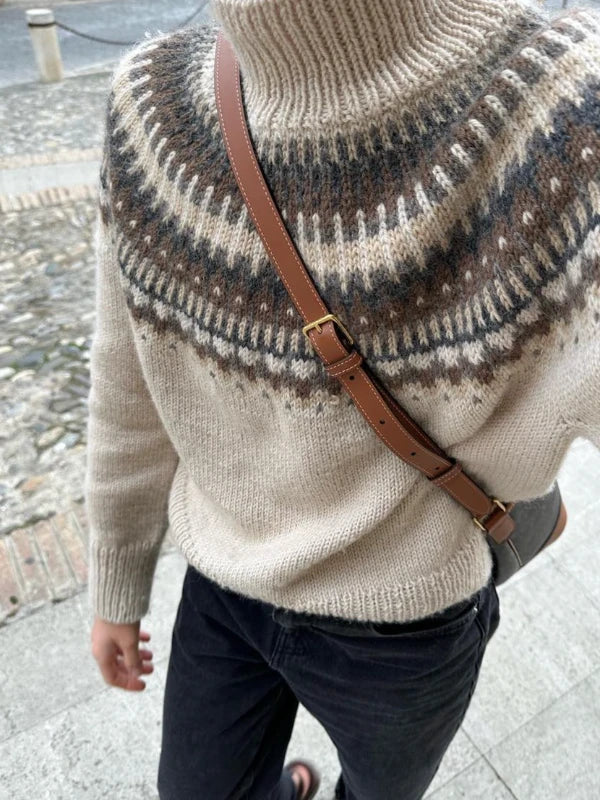 Wearing the PetiteKnit – Celeste Sweater neuleohje in a beige kirjoneule pattern with a cozy turtleneck and a brown crossbody strap, this person strolls outdoors along a stone pathway. The stylish knit design adds texture and charm, while their face remains unseen.