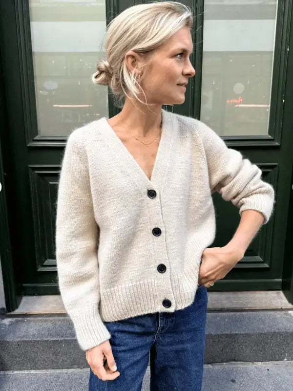 A woman with blonde hair in a low bun wears a cream-colored PetiteKnit - Champagne Cardigan neuleohje and blue jeans, standing outdoors by a dark green door, looking to the side.