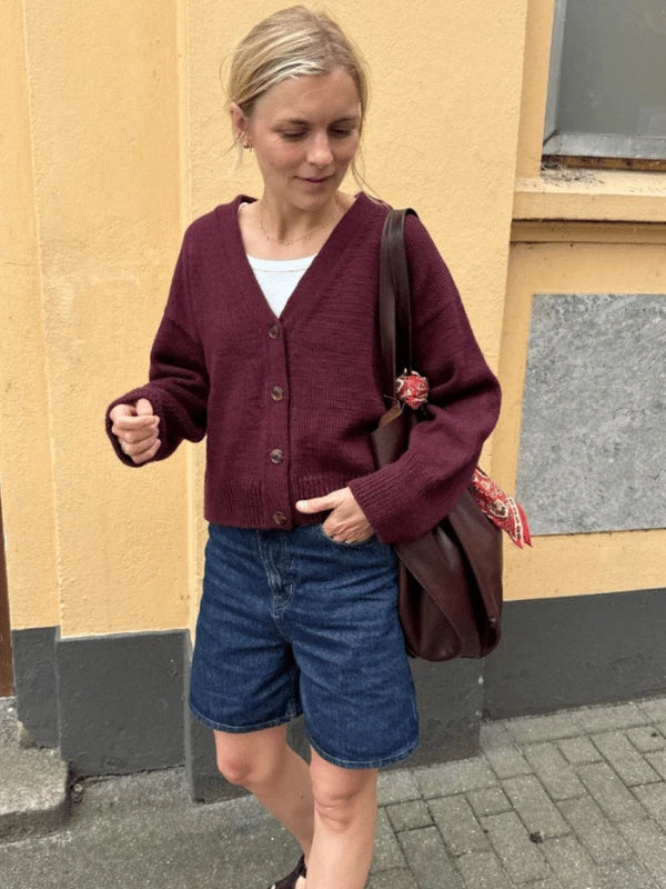 A woman stands outside by a yellow wall, smiling as she wears the PetiteKnit Scarlet Cardigan in burgundy over a white top with denim shorts, and carries a brown shoulder bag adorned with a red scarf.