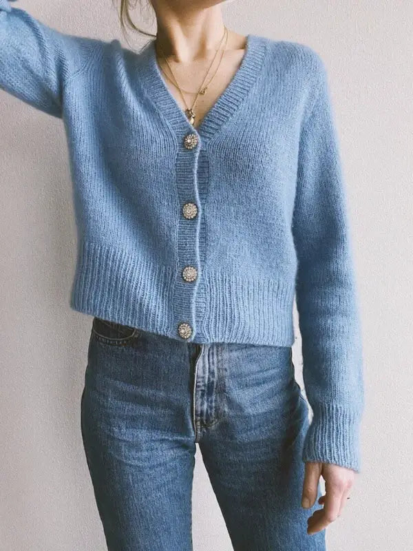 Wearing a light blue PetiteKnit - April Cardigan neuleohje with a V-neck and pearl-like buttons, paired with high-waisted blue jeans, the person stands before a plain light wall; their head appears mostly out of frame.