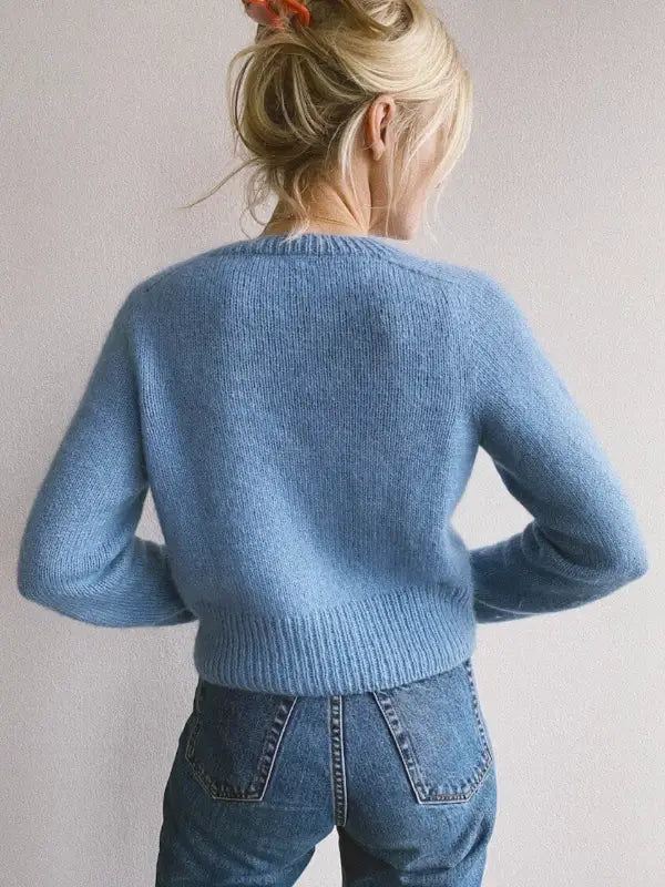 A person with blonde hair in a messy bun, secured with a red hair clip, wears the PetiteKnit - April Cardigan neuleohje and high-waisted blue jeans, standing with their back to the camera against a plain background.