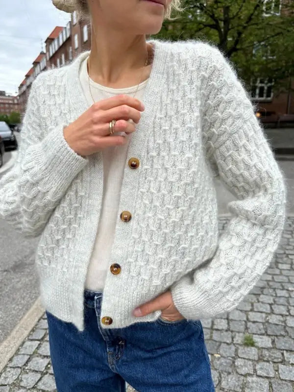 A person wears the PetiteKnit Jenny Jacket V-neck knit, a textured white cardigan with brown buttons, paired with a white top and blue jeans, standing outdoors with one hand in their pocket and the other touching their chest.