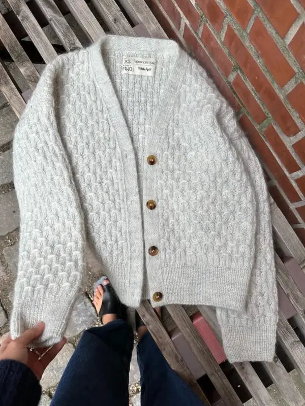 A PetiteKnit - Jenny Jacket V-neck neuleohje in textured light gray knit, featuring four brown buttons, is displayed flat on a wooden bench as someone in sandals and jeans holds a sleeve to highlight the knitting against a brick wall.