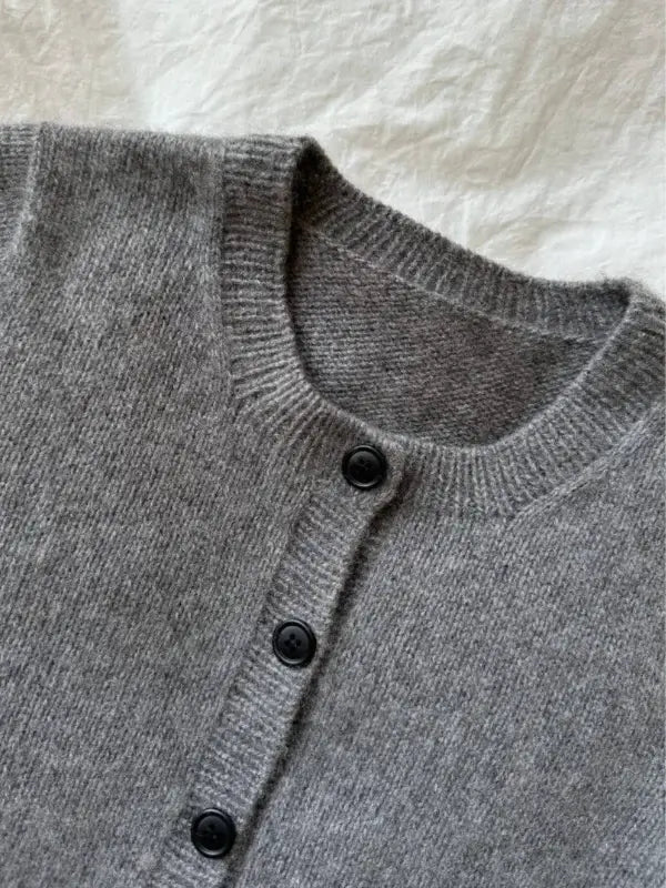 A close-up of the PetiteKnit Maggie Cardigan neuleohje, a gray knit sweater with black buttons, laid flat on a white textured surface.