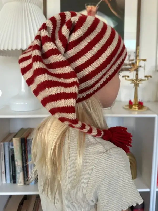A child with long blond hair wears the PetiteKnit tonttulakki neuleohje, Suomi—a red and white striped knit hat with a pointed end and pom-pom—while standing indoors near a white bookshelf and decorative items.